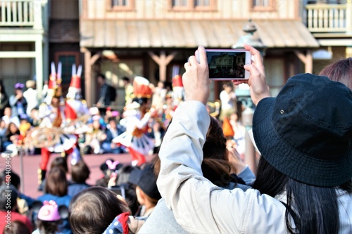 A girl in a black bucket hat stands with the crowd and takes a photo of a parade with her phone 