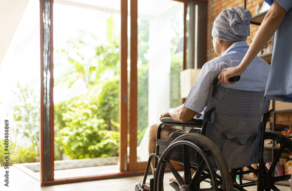 Attractive young Asian female nurse kneeling beside senior patient in wheelchair talking, smiling and cheering up in comfort at home.He survive from cancer .