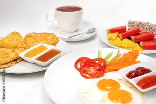 English breakfast. Scrambled eggs with tomato, green pepper and carrot on white plate, and pancakes with cup of tea and sliced fruit on white background