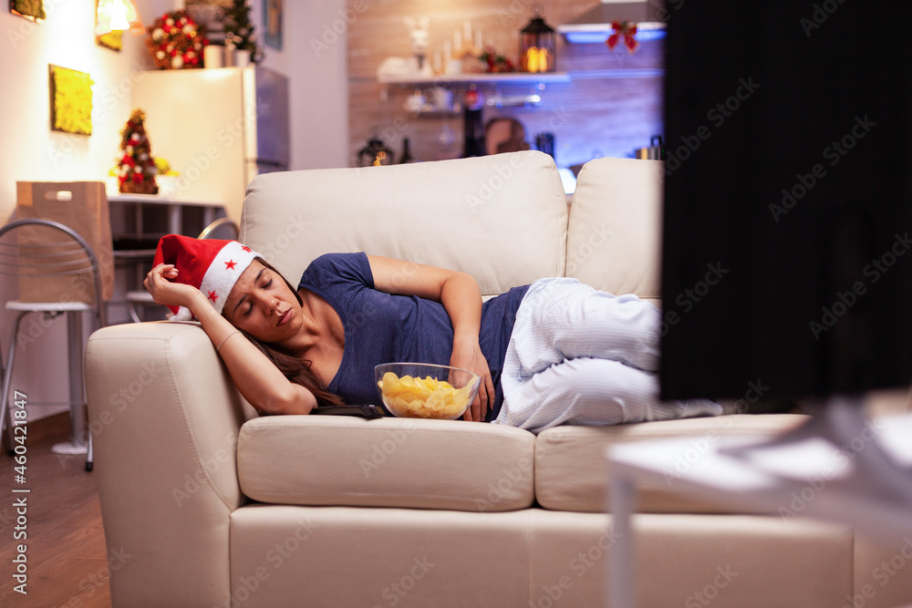 Obraz premium Tired woman falling asleep on sofa while watching xmas movie on television during christmastime enjoying winter season in x-mas decorated kitchen. Adult person celebrating christmas holiday
