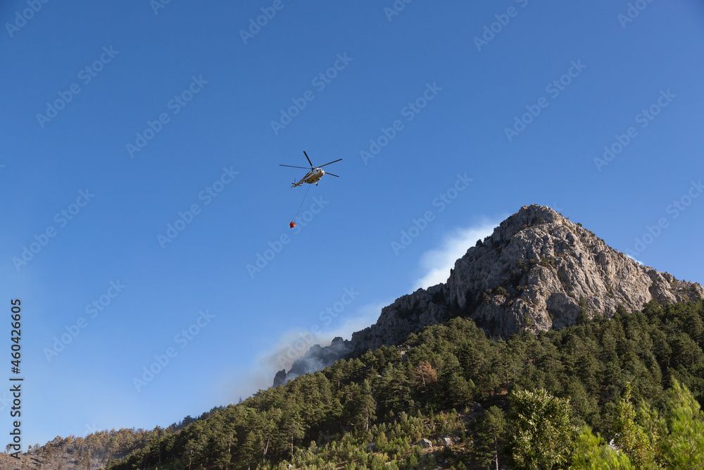 Wildfire helicopter goes to pour water on a fire on the slope of a steep, rocky hill.