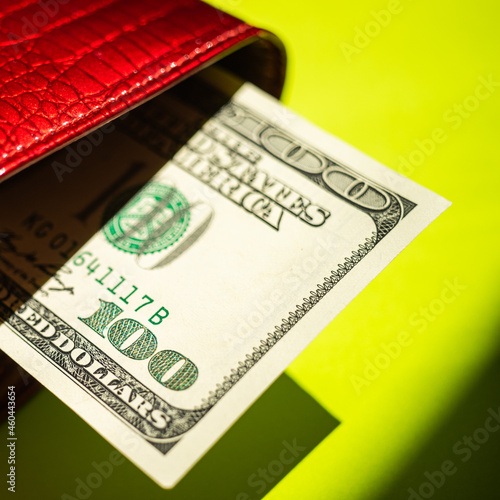 Hundred dollar bill sticking out of a red purse over sunny green table with strong shadows