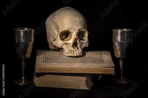 ludzka czaszka na książce i dwa metalowe kielichy na czarnym tle