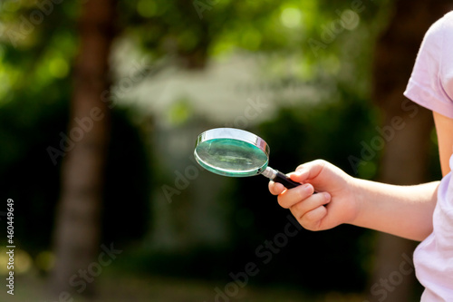 Young girl, little anonymous school age child holding a loupe, magnifying glass in hand, object closeup, blurred background, copy space. Clever kid, childhood exploration, education, curiosity concept