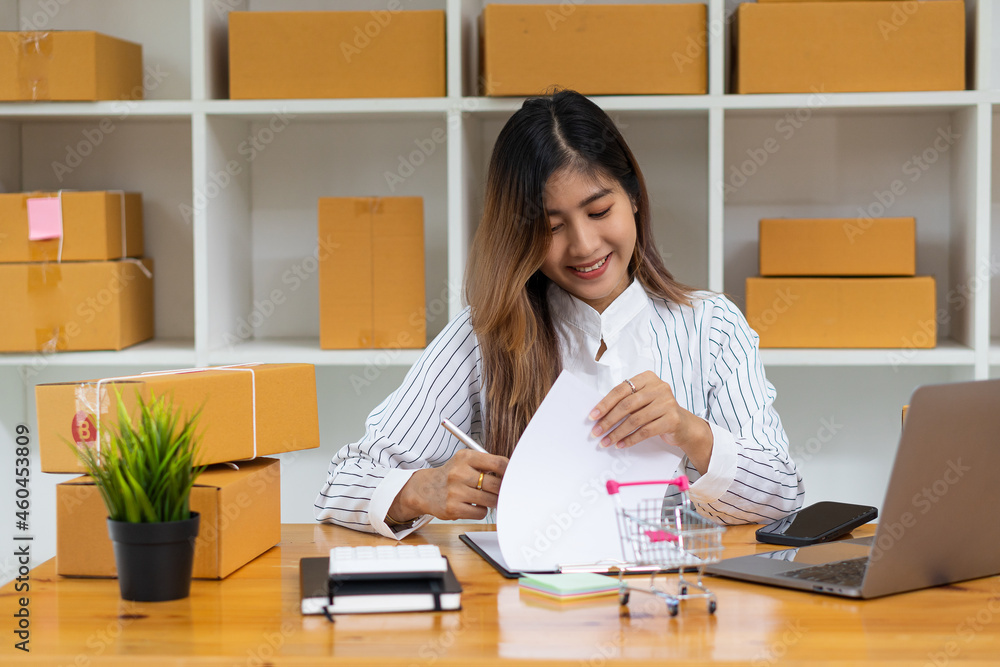 Beautiful Asian SME business owner online shopping working on laptop computer with parcel boxes on the desk at home - SME online business idea and box delivery.