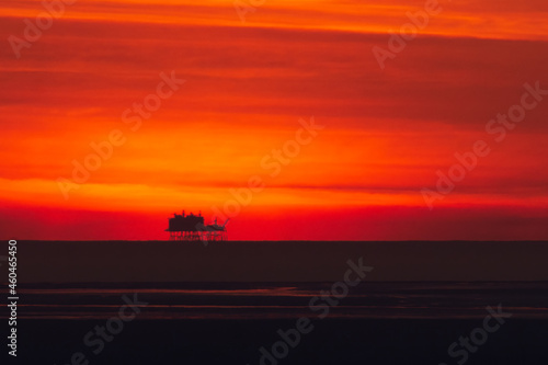 Distant oil rig or platform silhouette against orange sky at sunset. Theme of petrochemicals, sunset industry, industry, business © Callum