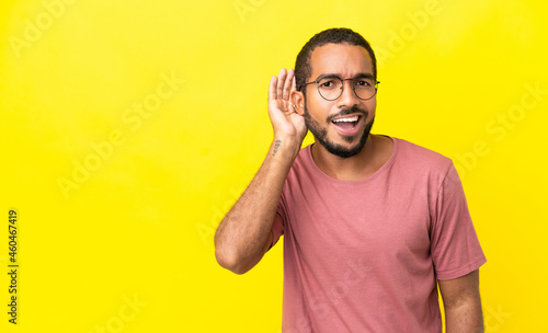 Young latin man isolated on yellow background listening to something by putting hand on the ear © luismolinero