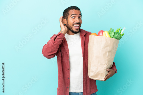 Young latin man holding a grocery shopping bag isolated on blue background listening to something by putting hand on the ear