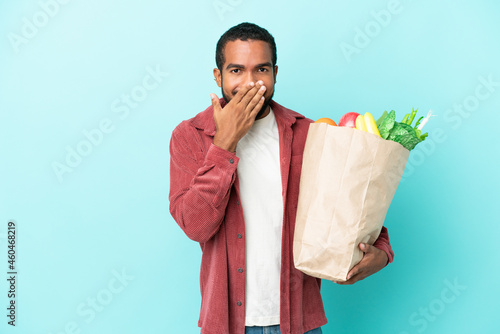 Young latin man holding a grocery shopping bag isolated on blue background happy and smiling covering mouth with hand