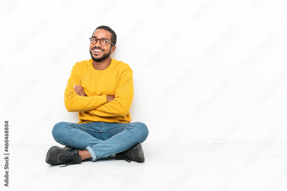 Young Ecuadorian man sitting on the floor isolated on white wall with arms crossed and happy Young Ecuadorian man sitting on the floor isolated on white wall with arms crossed and happy