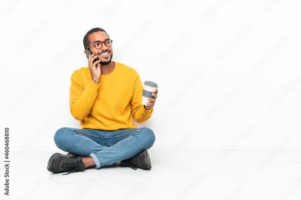 Young Ecuadorian man sitting on the floor isolated on white wall holding coffee to take away and a mobile Young Ecuadorian man sitting on the floor isolated on white wall holding coffee to take away and a mobile