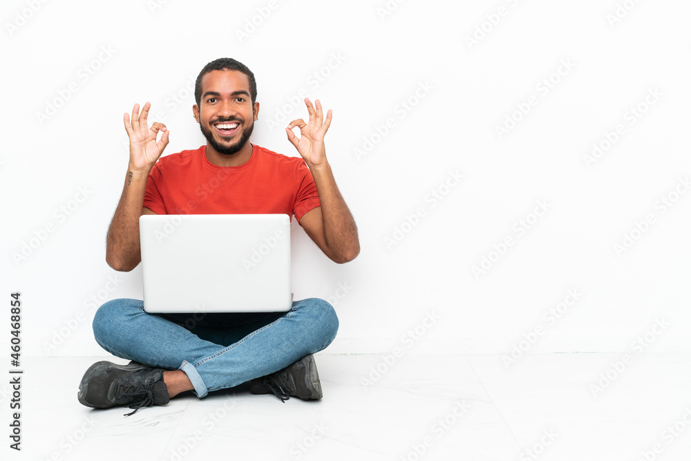 Young Ecuadorian man with a laptop sitting on the floor isolated on white background showing an ok sign with fingers Young Ecuadorian man with a laptop sitting on the floor isolated on white background showing an ok sign with fingers