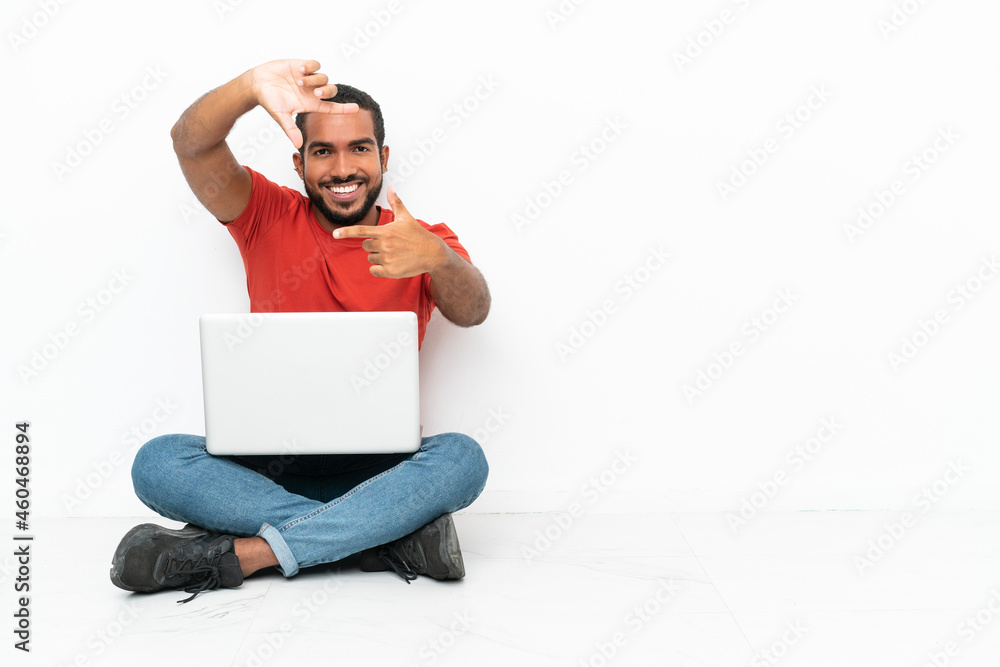 Young Ecuadorian man with a laptop sitting on the floor isolated on white background focusing face. Framing symbol Young Ecuadorian man with a laptop sitting on the floor isolated on white background focusing face. Framing symbol