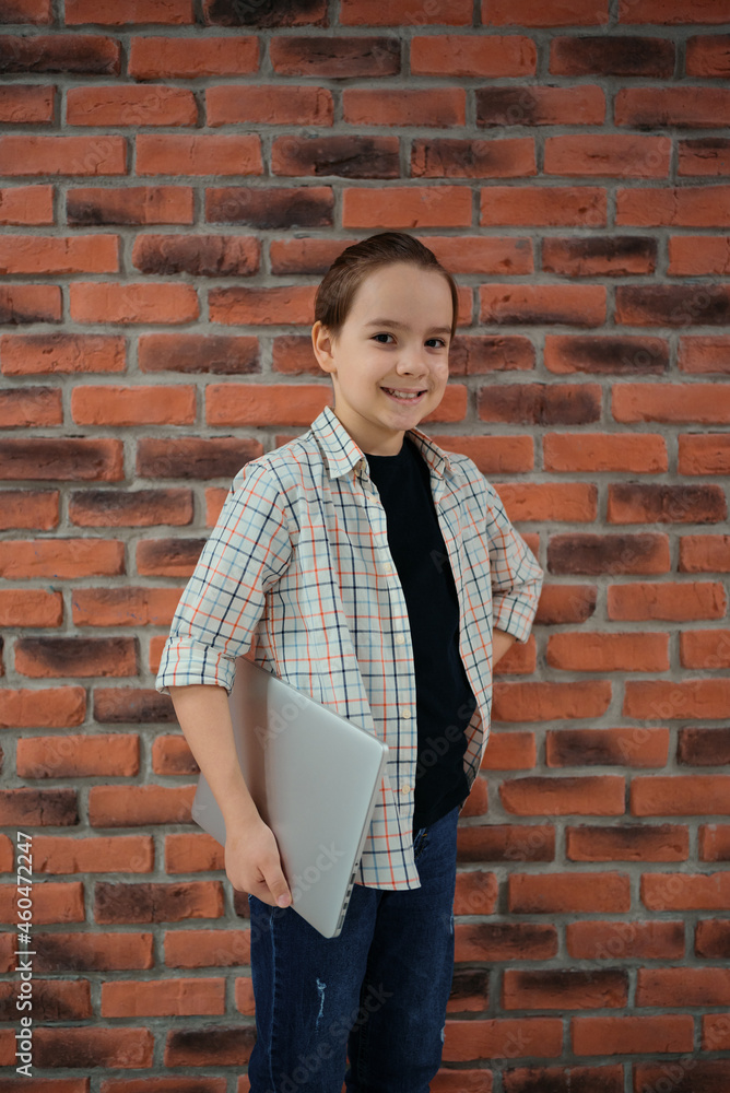 Indoor portrait of Caucasian boy dressed in shirt standing against brick wall holding laptop. Indoor portrait of Caucasian boy dressed in shirt standing against brick wall holding laptop.