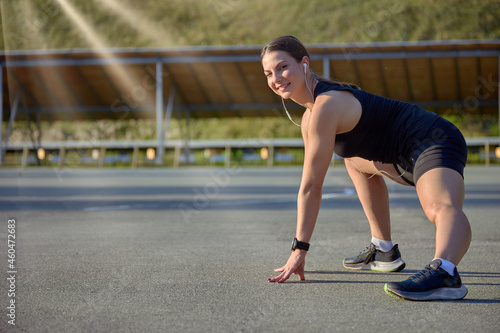 Portrait of a fit and athletic young woman in headphones and black sportswear doing stretching at the stadium in the rays of sunlight. Concept of a healthy lifestyle in sports.