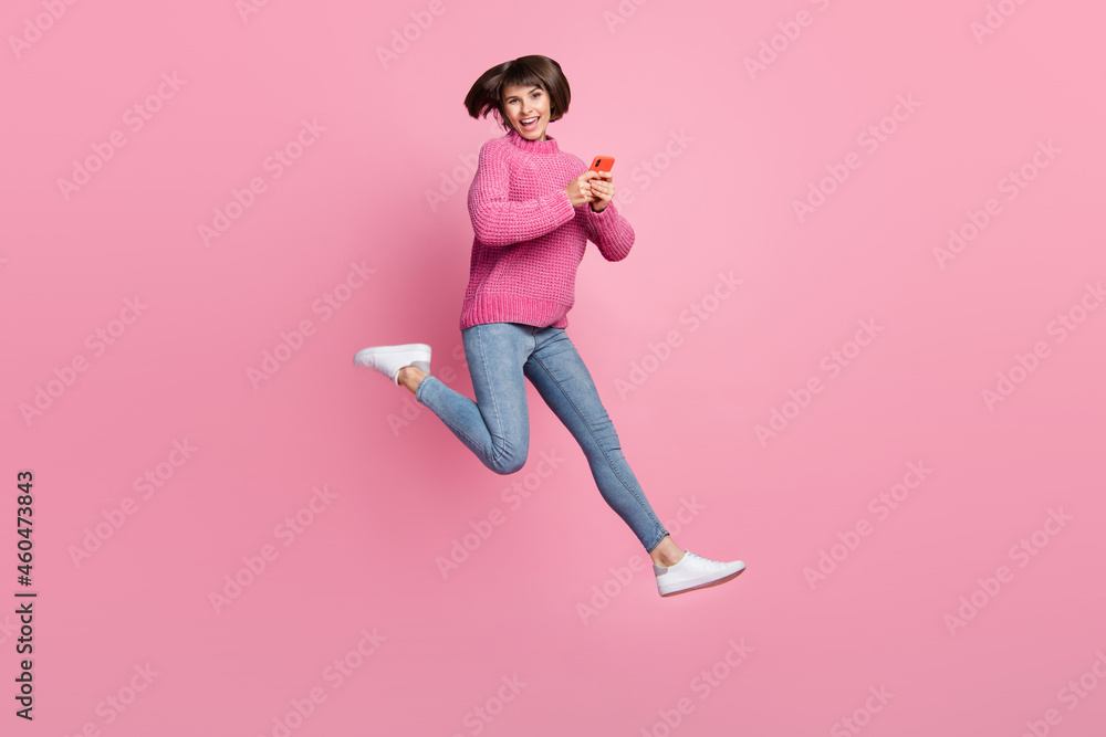 Full length photo of cheerful happy young woman jump up hold hands phone smile isolated on pink color background Full length photo of cheerful happy young woman jump up hold hands phone smile isolated on pink color background
