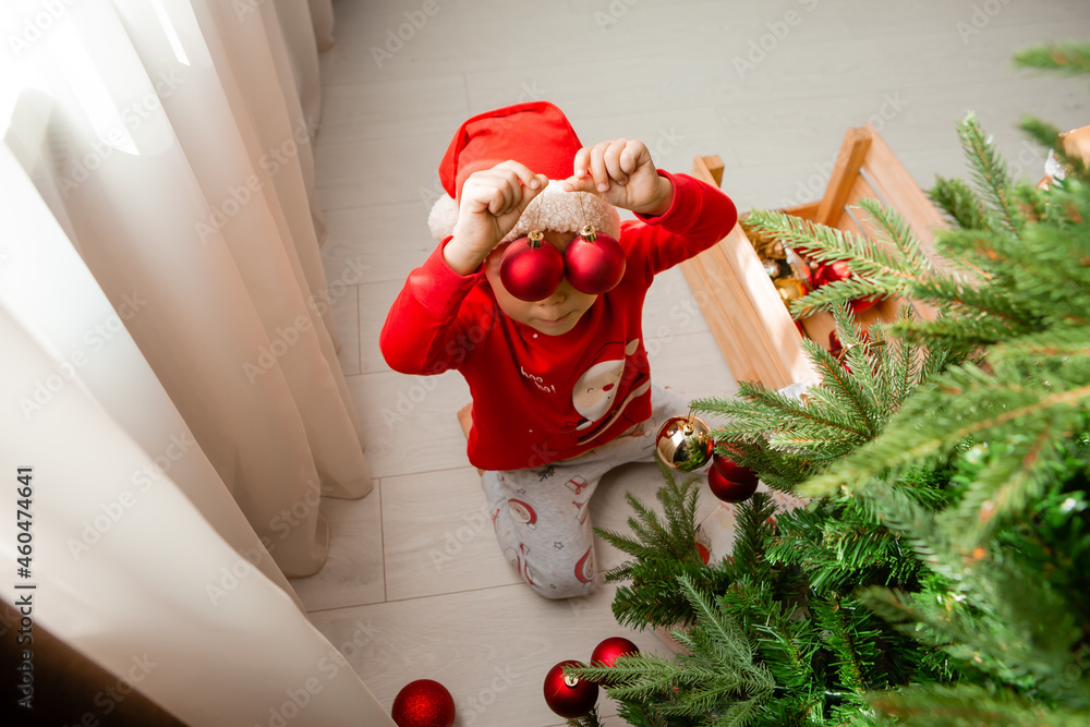 Fototapeta premium little boy in red pajamas decorates the Christmas tree with toys and balls. Cute kid preparing home for xmas celebration. winter New Year's concept. space for the text. High quality photo