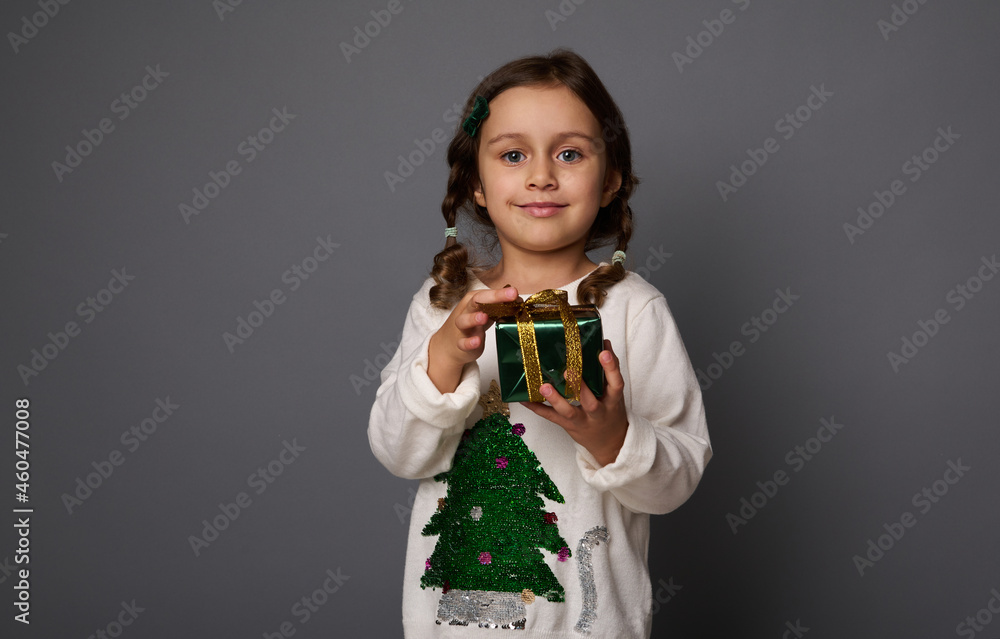 Beautiful little girl in white sweater with a Christmas tree, poses against gray background, holding a Christmas gift in green glitter wrapping paper and golden ribbon, cute smiles looking at camera Beautiful little girl in white sweater with a Christmas tree, poses against gray background, holding a Christmas gift in green glitter wrapping paper and golden ribbon, cute smiles looking at camera