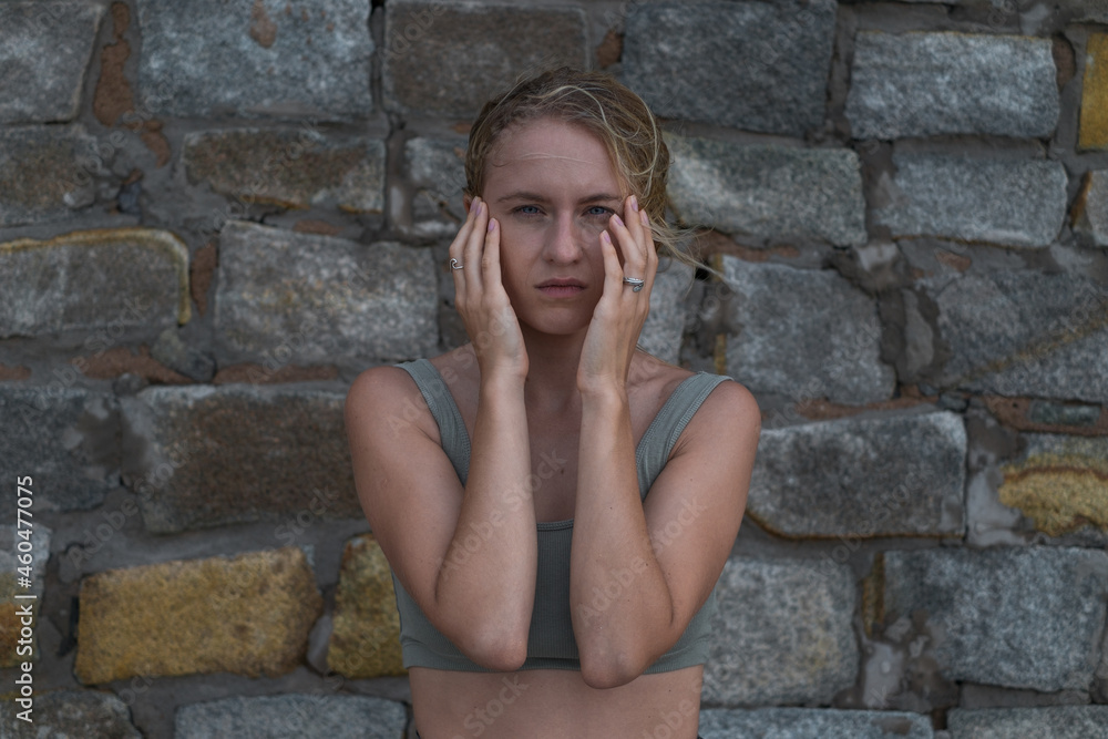 Sad stressful face of young caucasian blonde woman in crop top on the stone background. Young woman crying and using hands to cover face.