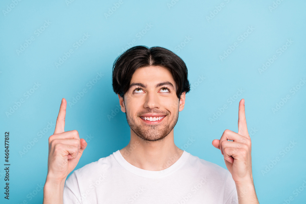 Photo of funny young brunet guy point look up empty space wear white t-shirt isolated on blue color background Photo of funny young brunet guy point look up empty space wear white t-shirt isolated on blue color background