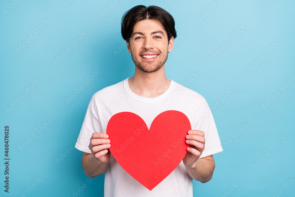 Photo of flirty young brunet guy hold heart wear white t-shirt isolated on blue color background Photo of flirty young brunet guy hold heart wear white t-shirt isolated on blue color background