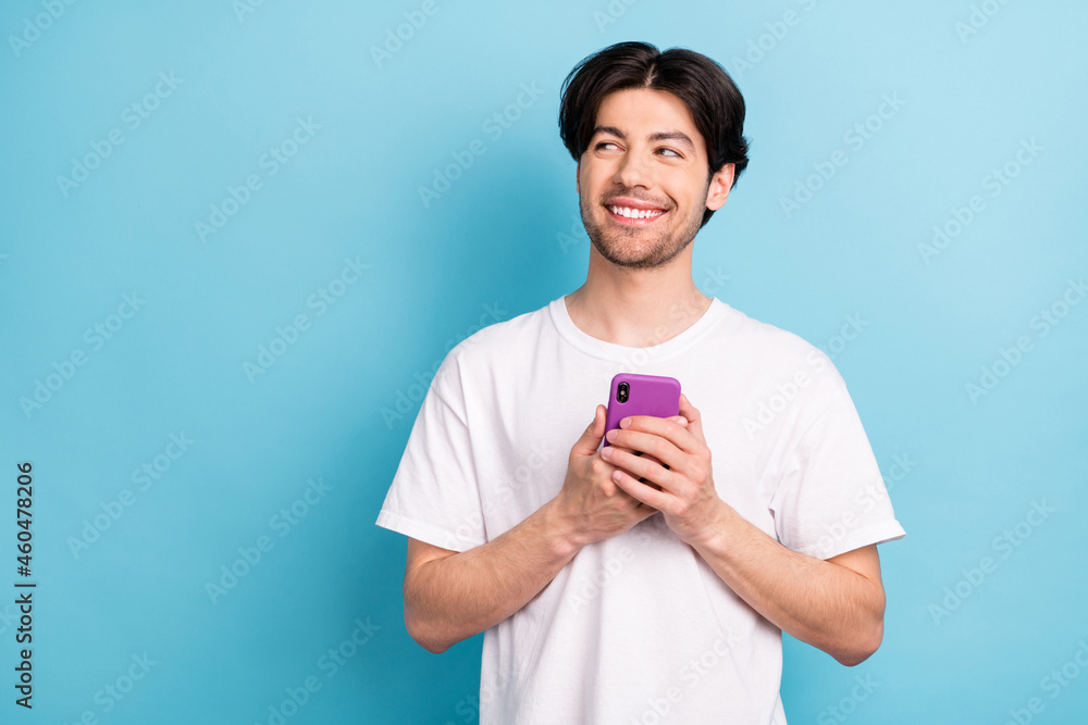 Photo of funny millennial brunet guy hold telephone look empty space wear white t-shirt isolated on blue color background Photo of funny millennial brunet guy hold telephone look empty space wear white t-shirt isolated on blue color background