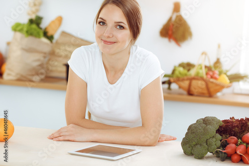 An attractive young woman looking for a new recipe for a delicious salad mix while sitting at the table in sunny kitchen. Tablet pc is the best cookbook