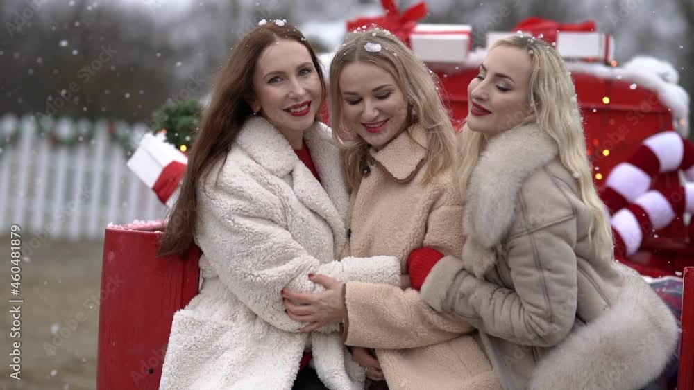 Three young women in winter coats sitting in red Christmas car with New Year decor, hugging, posing to the camera. Holiday concept. Slow motion. Three young women in winter coats sitting in red Christmas car with New Year decor, hugging, posing to the camera. Holiday concept. Slow motion.