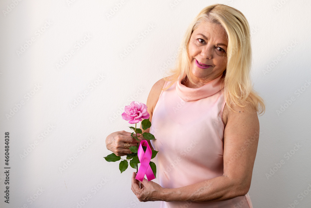 October pink, blonde lady in light pink blouse, holding beside her body pink flower with ribbon symbol of October pink campaign, light background, looking and smiling at photo