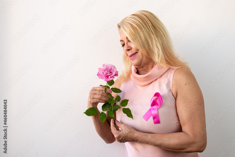 Pink October, blonde lady in a light pink blouse with ribbon, symbol of the Pink October campaign, looking sideways with affection and smiling at pink flower, light background