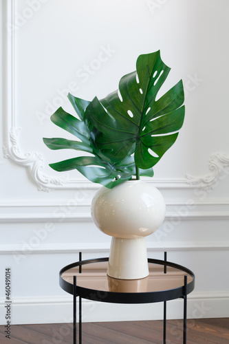 White ceramic vase with monstera leaves on a mirrored coffee table against a white wall with stucco molding. Modern interior details. Stylish room decoration. Vertical image
