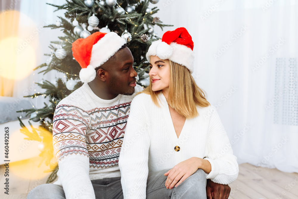 Obraz premium Portrait of a couple in love in santa hats sitting by the tree. Glare of the yellow garland. Black male hugs a Caucasian woman