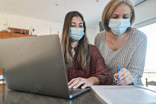 Twin sisters and their mother wearing protective mask and working a elearning lesson on  a laptop at home during lockdown due to covid-19