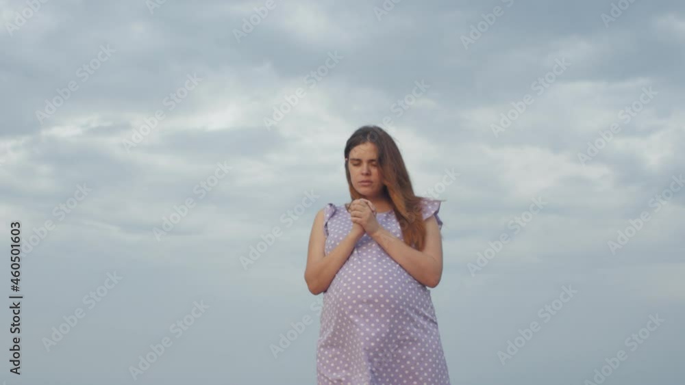 young pregnant woman folded her hands in pray to God standing alone in field against of cloudy sky, future mother walking on nature young pregnant woman folded her hands in pray to God standing alone in field against of cloudy sky, future mother walking on nature