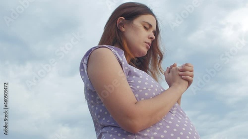 young pregnant woman folded her hands in pray to God standing alone in field against of cloudy sky, future mother walking on nature