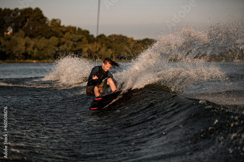 Strong athletic male wakeboarder holds rope and energetically riding wakeboard on splashing river water.