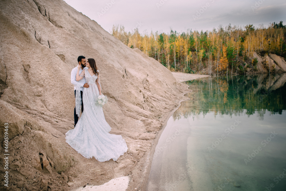 Newlyweds are standing near a beautiful lake. Middle Eastern groom and Caucasian bride embrace on the beach.