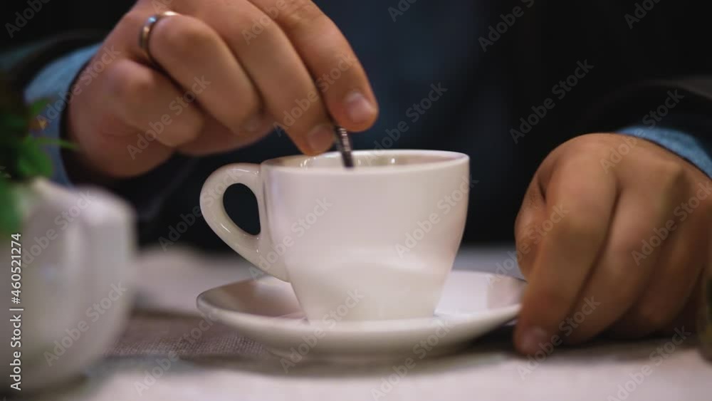 Man in outerwear mixes tea or coffee with teaspoon and puts spoon on saucer and takes cup with hand. Person drinks hot drink to warm up and enjoy and relax. Cup with saucer on table on terrace Man in outerwear mixes tea or coffee with teaspoon and puts spoon on saucer and takes cup with hand. Person drinks hot drink to warm up and enjoy and relax. Cup with saucer on table on terrace