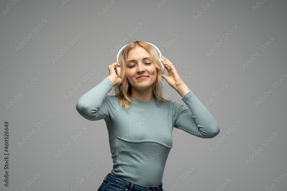 Beautiful attractive young blond woman wearing blue t-shirt and glasses in white headphones listening music and smiling on grey background in studio. Relaxing and enjoying. Lifestyle. Beautiful attractive young blond woman wearing blue t-shirt and glasses in white headphones listening music and smiling on grey background in studio. Relaxing and enjoying. Lifestyle.