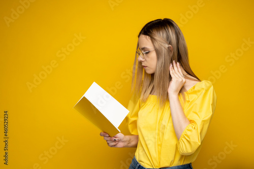 Beautiful smart young girl holding and reading book isolated on the yellow background. Portrait of attractive woman in a yellow blouse and wearing glasses reading book. Education, studying, knowledge.