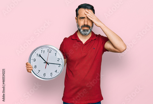 Middle aged man with beard holding big clock stressed and frustrated with hand on head, surprised and angry face