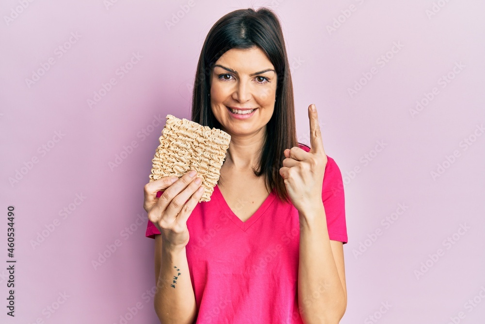 Young hispanic woman holding uncooked noodles smiling with an idea or question pointing finger with happy face, number one Young hispanic woman holding uncooked noodles smiling with an idea or question pointing finger with happy face, number one