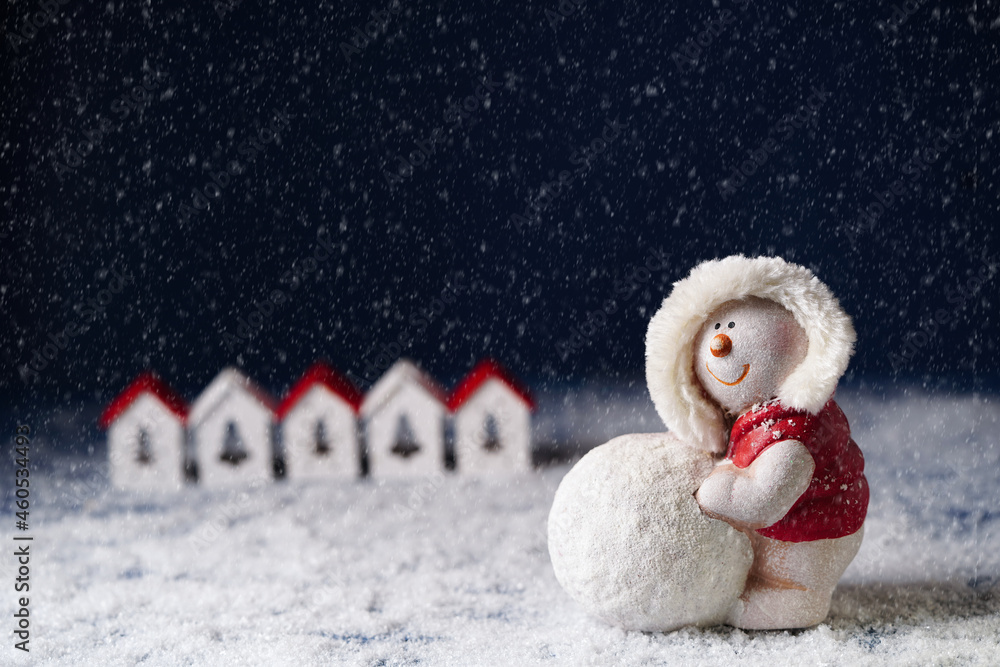 a snowman rolls a snowball on the background of small houses on a dark blue background, a place for congratulations on Christmas and New Year. a snowman rolls a snowball on the background of small houses on a dark blue background, a place for congratulations on Christmas and New Year.