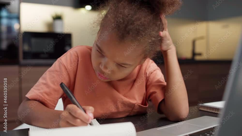 Intelligent biracial little girl doing homework at home. Kid writing in notepad and typing on laptop pc sitting at table. Distance education and online learning at quarantine concept Intelligent biracial little girl doing homework at home. Kid writing in notepad and typing on laptop pc sitting at table. Distance education and online learning at quarantine concept