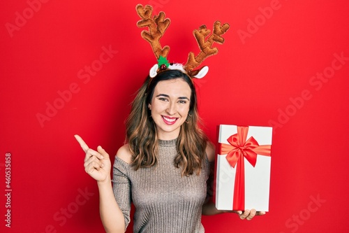 Young hispanic girl wearing deer christmas hat holding gift smiling happy pointing with hand and finger to the side