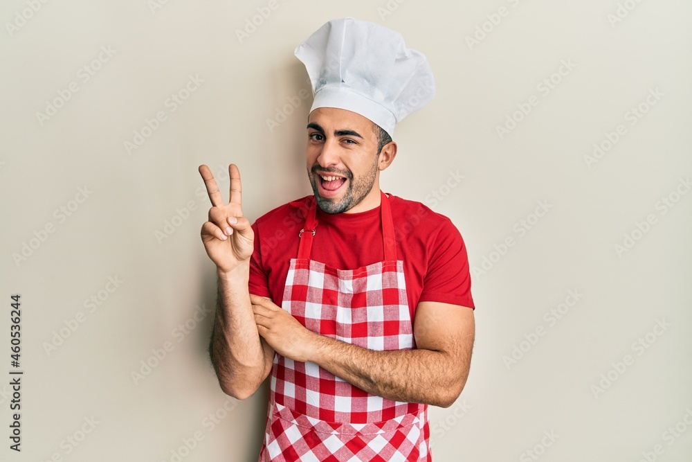 Young hispanic man wearing baker uniform smiling with happy face winking at the camera doing victory sign. number two. Young hispanic man wearing baker uniform smiling with happy face winking at the camera doing victory sign. number two.