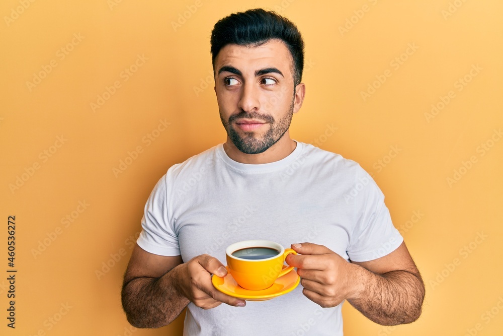 Young hispanic man drinking a cup of coffee smiling looking to the side and staring away thinking.