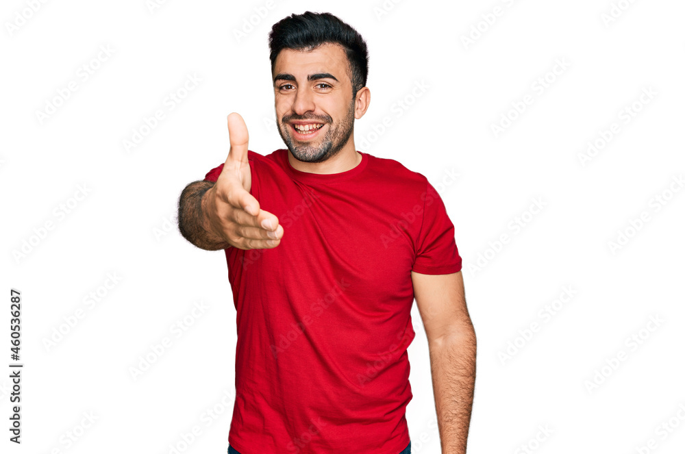 Hispanic man with beard wearing casual red t shirt smiling friendly offering handshake as greeting and welcoming. successful business. Hispanic man with beard wearing casual red t shirt smiling friendly offering handshake as greeting and welcoming. successful business.