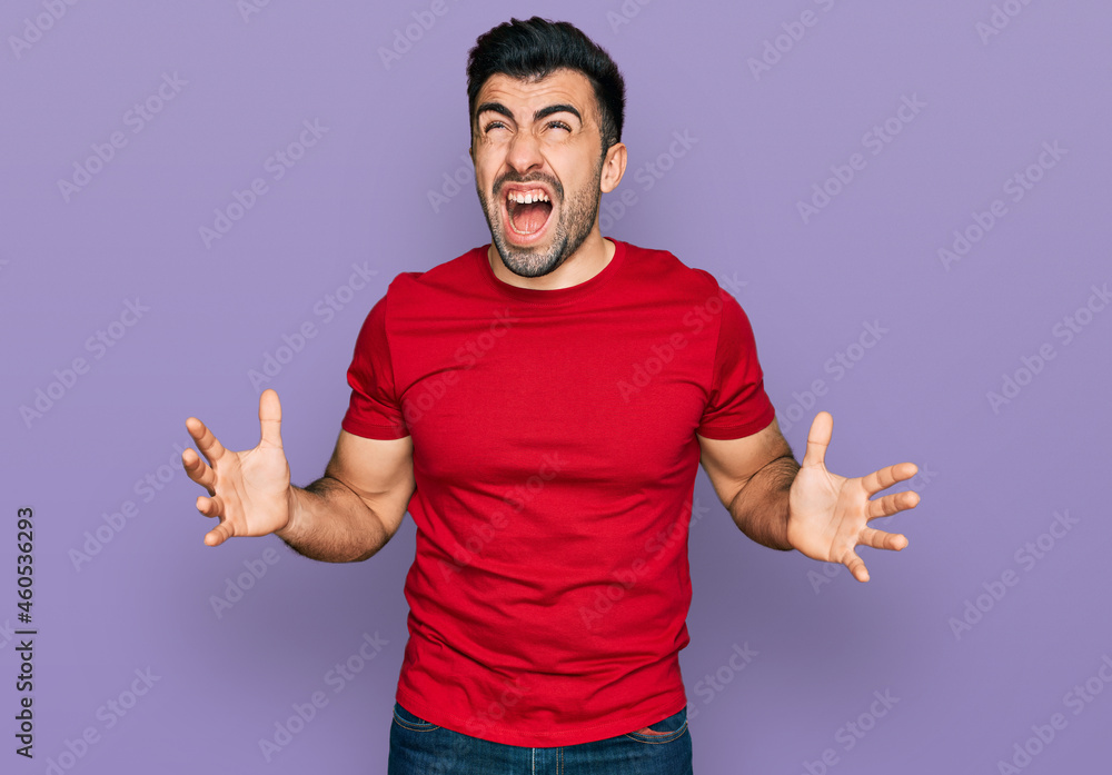 Hispanic man with beard wearing casual red t shirt crazy and mad shouting and yelling with aggressive expression and arms raised. frustration concept. Hispanic man with beard wearing casual red t shirt crazy and mad shouting and yelling with aggressive expression and arms raised. frustration concept.