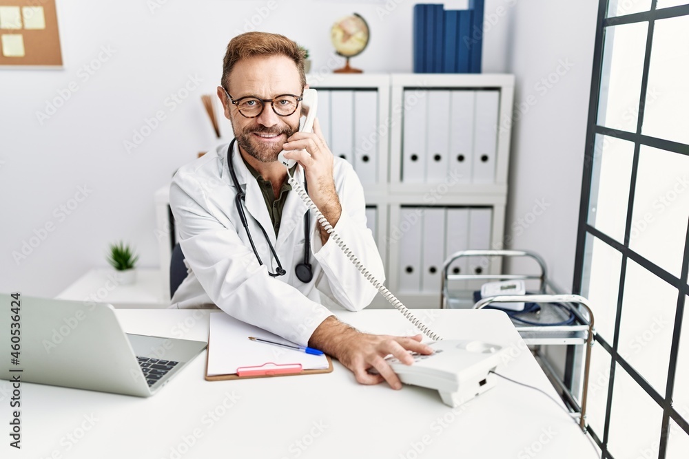 Middle age hispanic man wearing doctor uniform talking on the telephone at clinic